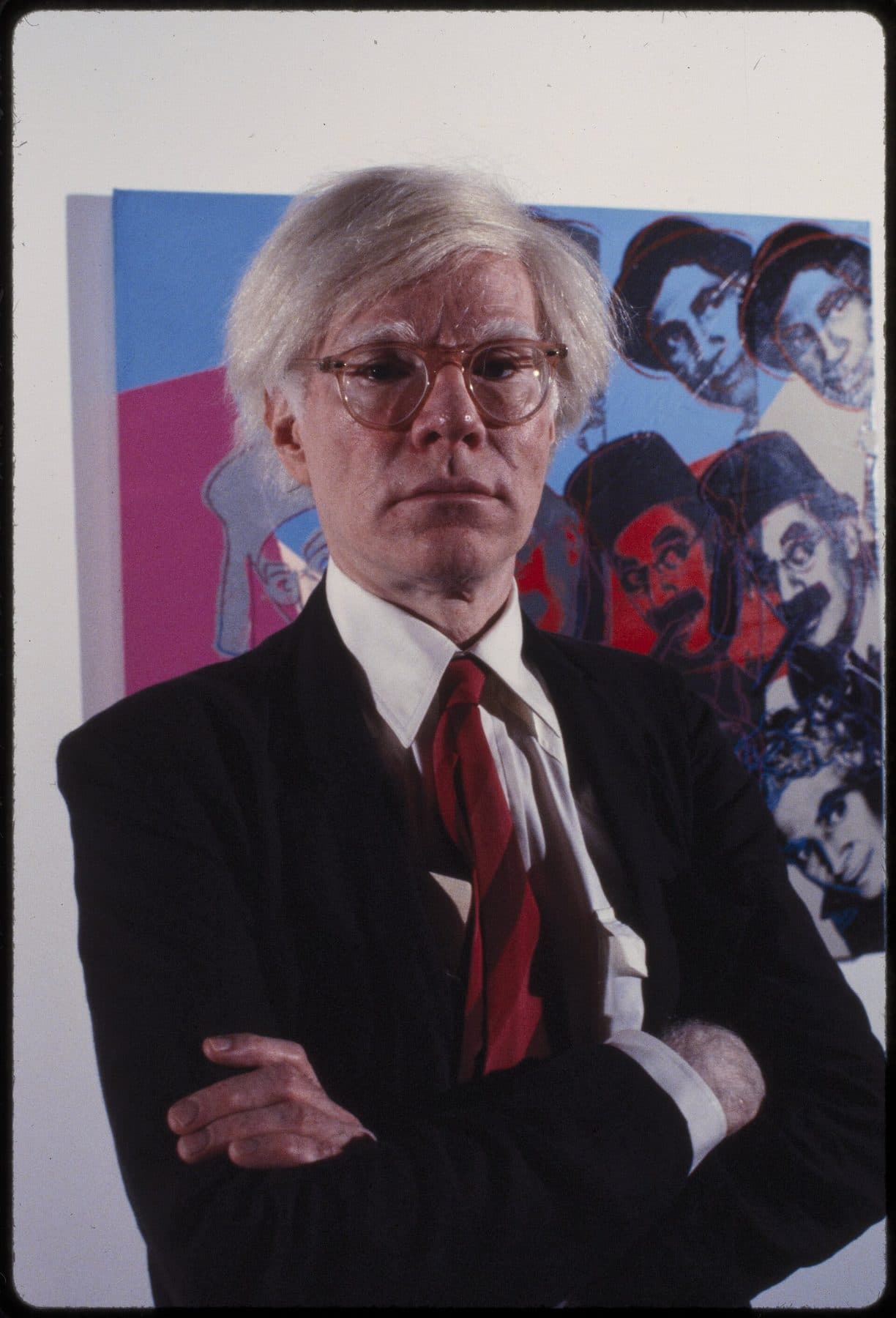 Andy Warhol, arms folded, in a dark suit and red tie, in front of his screenprint portraits at the Jewish Museum in New York, 1980.