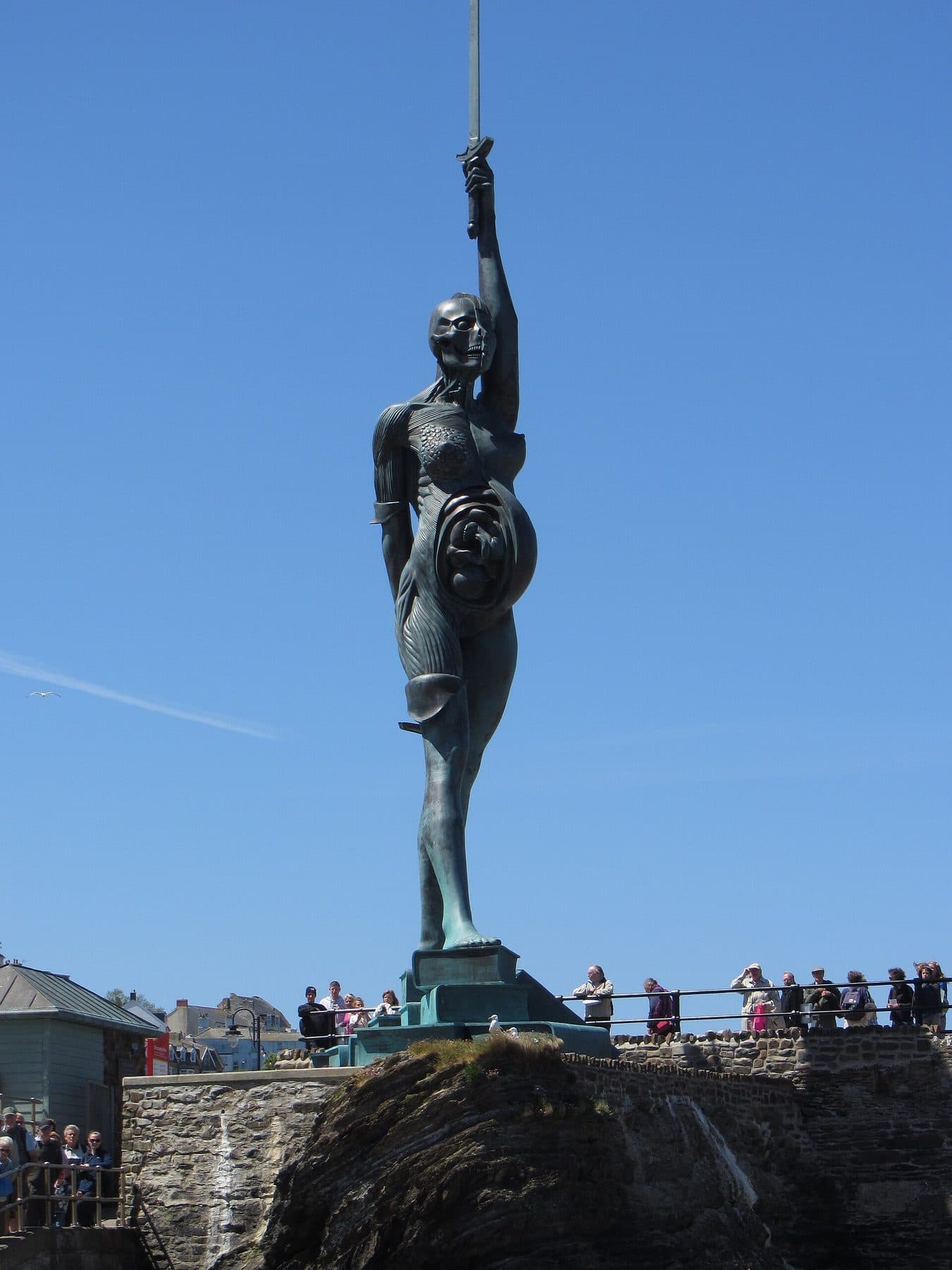 Damien Hirst's giant bronze Verity, a pregnant woman holding a sword aloft, standing on the harbour wall at Ilfracombe against a blue sky.