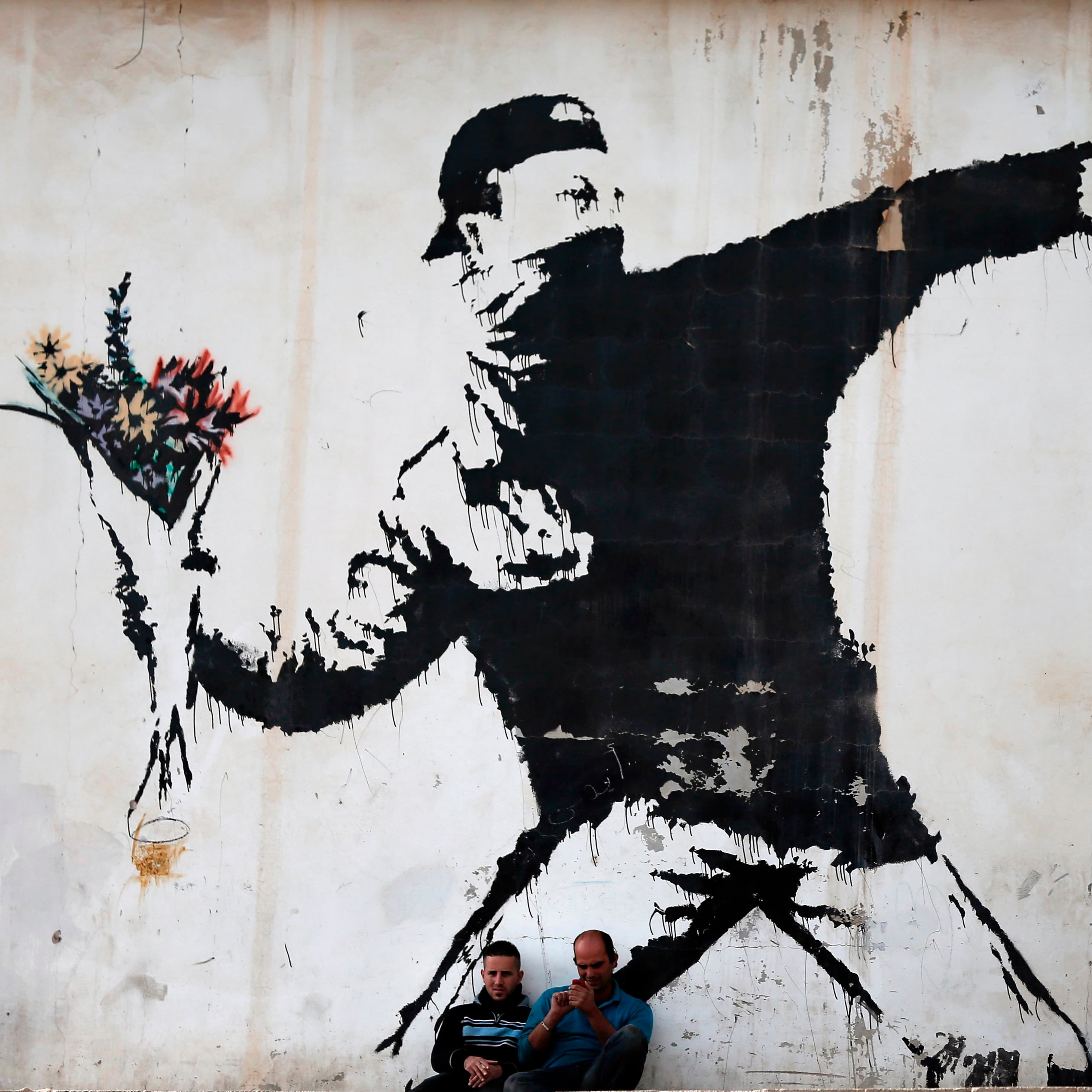 Banksy's Flower Thrower stencilled on a wall in the West Bank: a masked figure winding up to throw, holding a bouquet of flowers instead of a missile, with two men sitting on the ground beneath it