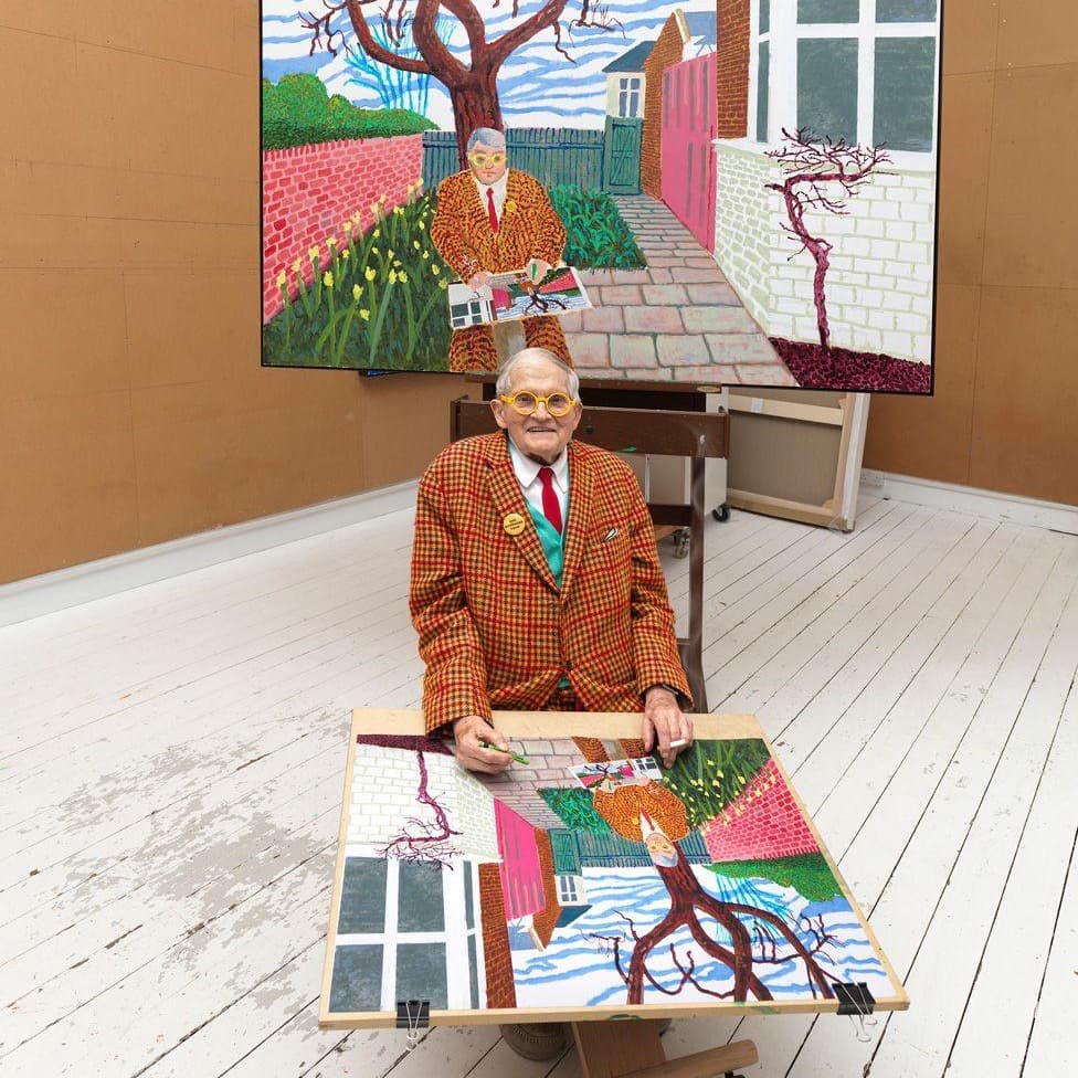 David Hockney in a checked jacket and yellow round glasses, seated at a drawing board in his studio working on a self-portrait, with the finished painting on an easel behind him
