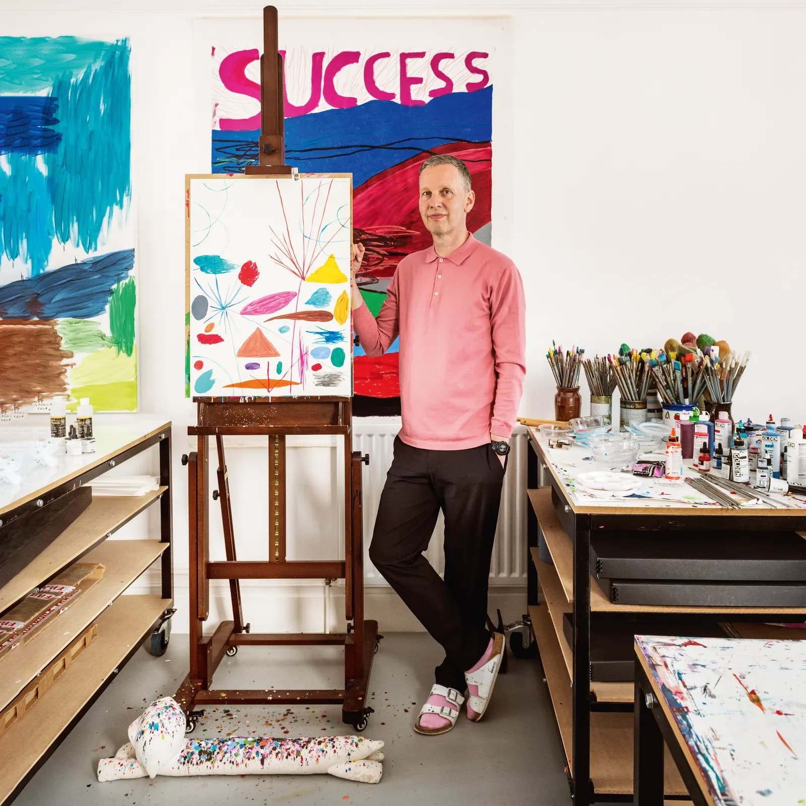 David Shrigley in his studio in a pink polo shirt, leaning against an easel holding a drawing of scattered coloured marks, with a large painting reading SUCCESS on the wall behind him