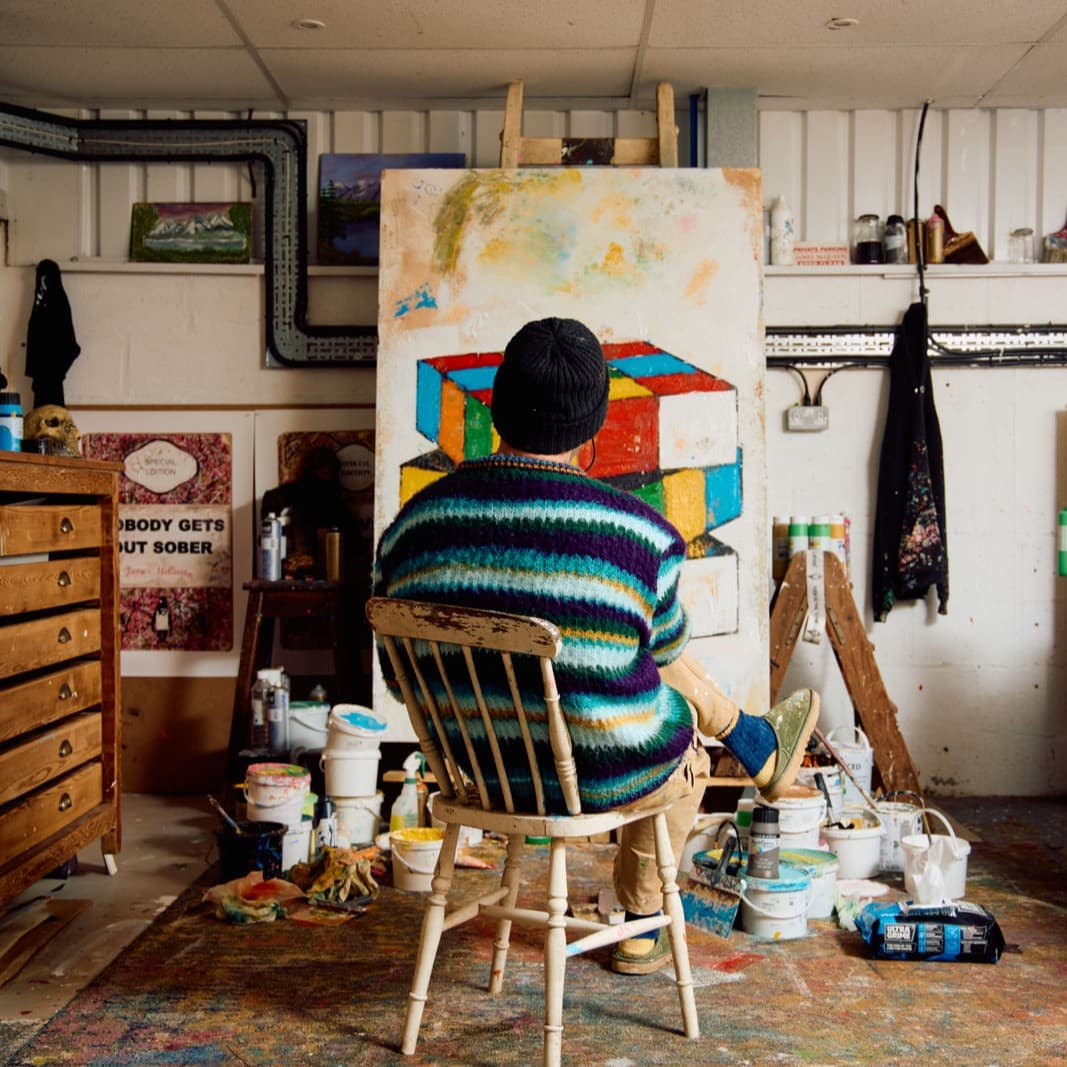 James McQueen seated from behind in his studio, working on a large canvas of interlocking coloured blocks, with paint pots across the floor and one of his own printed label works propped against the wall