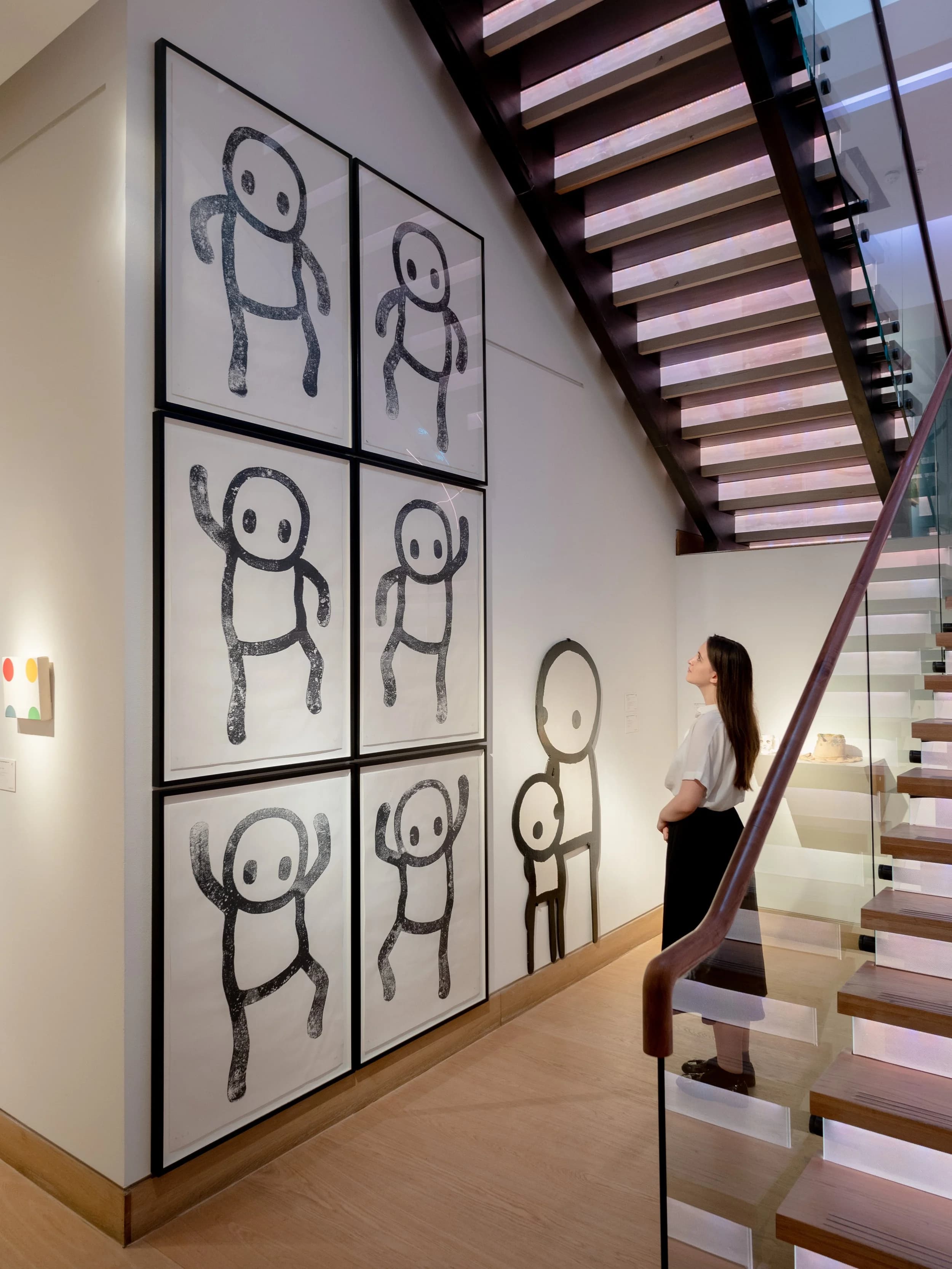 Six framed Stik works on paper hung in a grid beside a painted Stik figure, in a gallery stairwell.