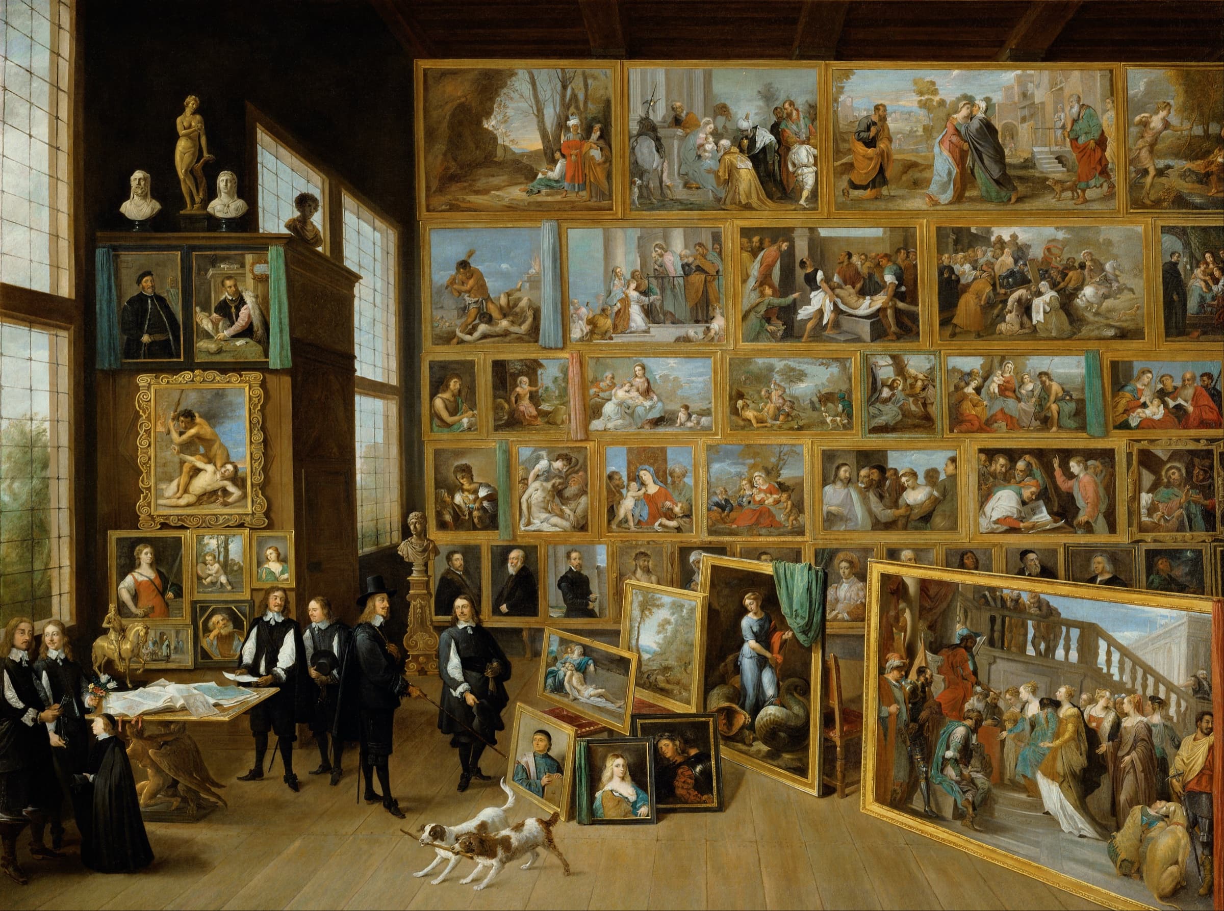 A seventeenth-century painting of a picture gallery: the walls hung frame to frame with paintings, more stacked against them on the floor, and the Archduke and his courtiers looking them over while two dogs play