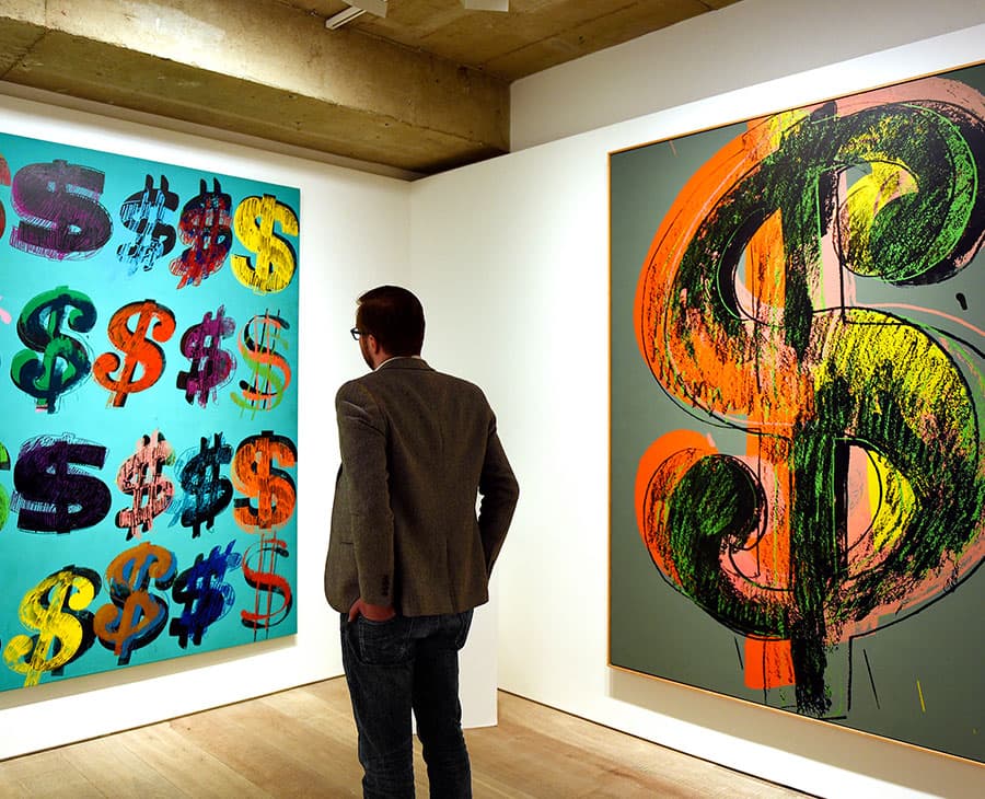 Two Andy Warhol Dollar Sign paintings hanging in a gallery, with a visitor standing between them