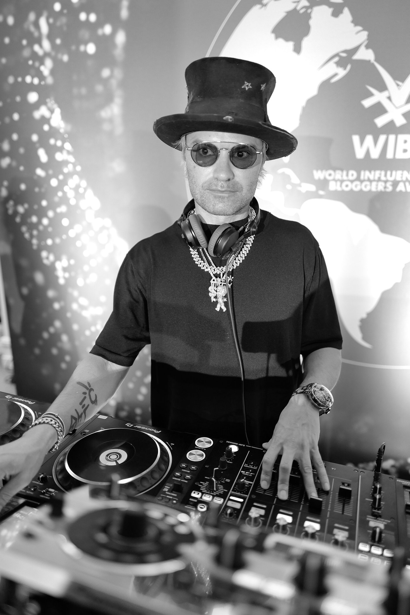 Alec Monopoly in a top hat, round dark glasses and headphones, with a chain round his neck.
