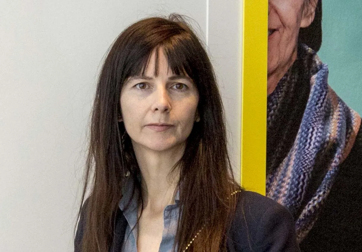 Gillian Wearing in a navy jacket and grey shirt, standing in front of large photographic portraits