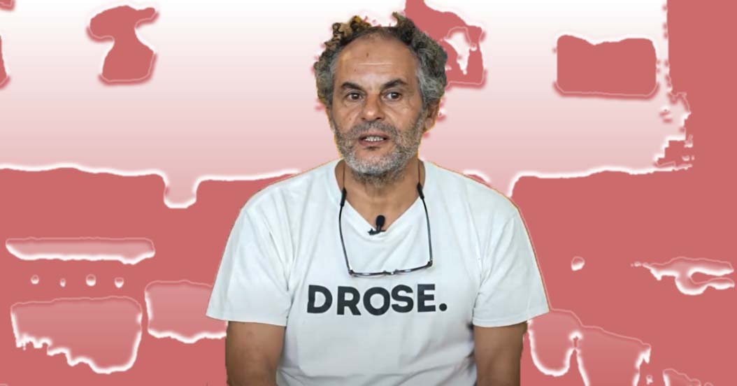 Hassan Hajjaj in a white T-shirt, being interviewed against a red and pink backdrop.