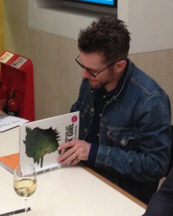 Jamie Hewlett in glasses and a denim jacket, signing books at a table.