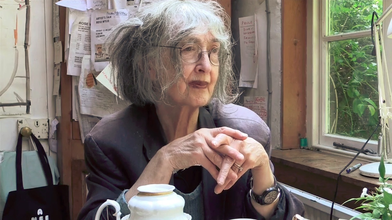 Rose Wylie seated by a window in her studio, hands clasped, with notes pinned to the wall behind her