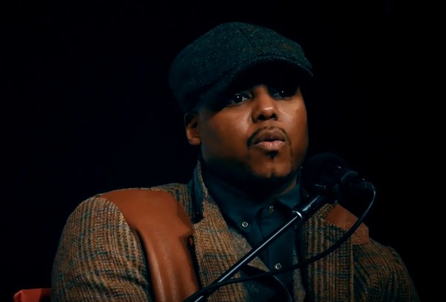 Titus Kaphar in a cap and tweed jacket, speaking into a microphone.