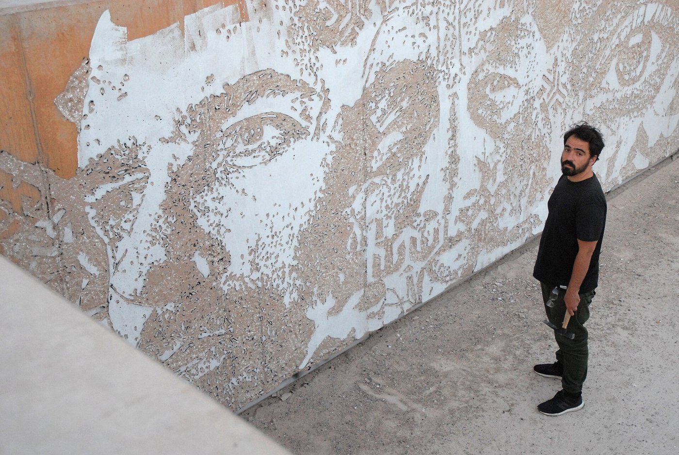 Vhils standing beside a huge face carved into a white wall.
