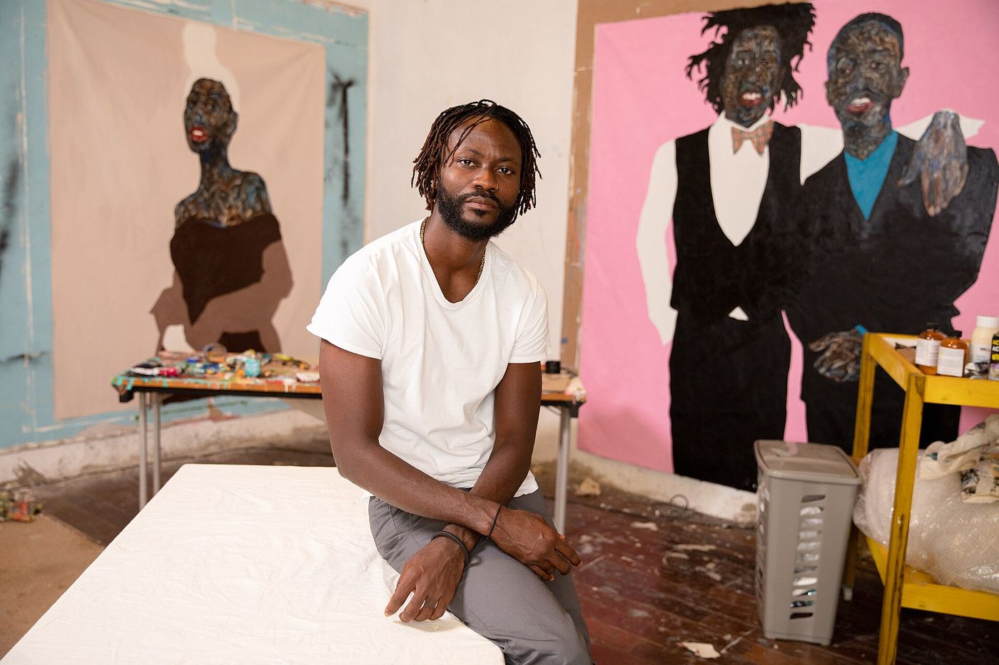 Amoako Boafo in a white T-shirt, seated in his studio in front of two of his large portrait paintings
