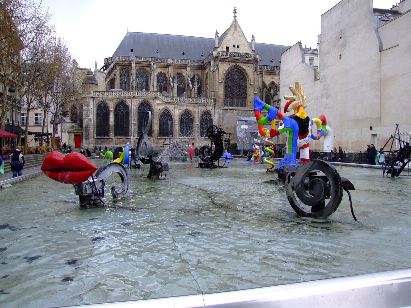 The Stravinsky Fountain in Paris: black machine sculptures and brightly painted figures, among them a pair of red lips, spraying water in a shallow pool beside a Gothic church.