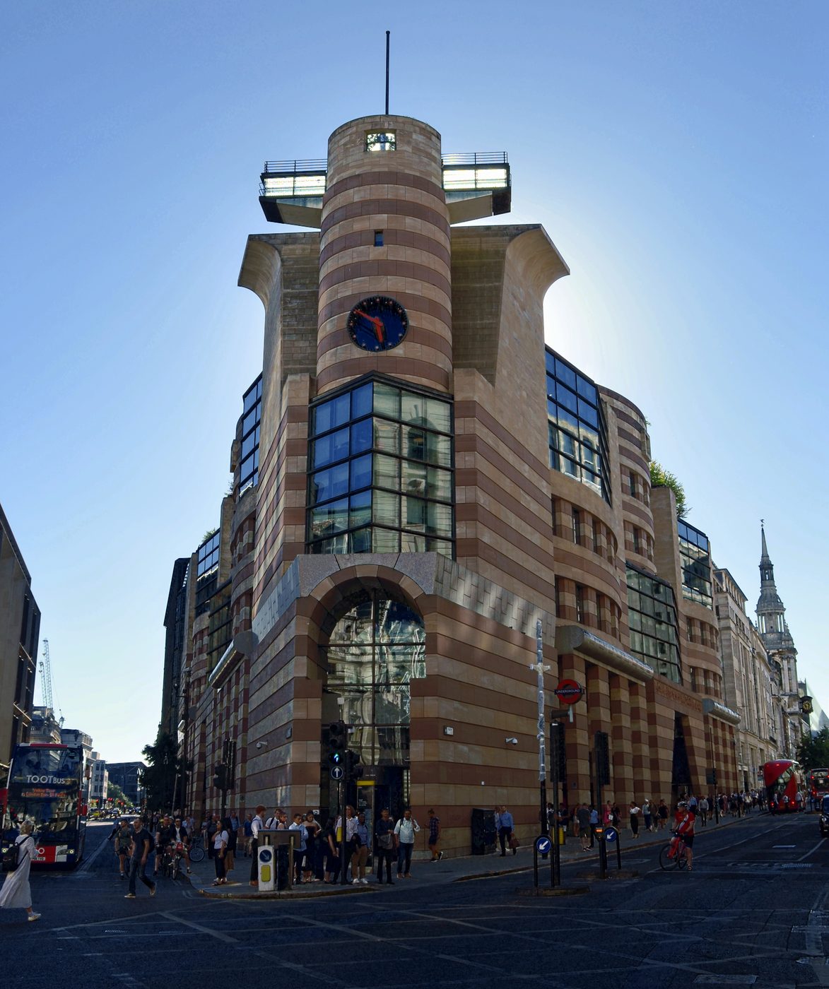 No 1 Poultry in the City of London: a building banded in pink and yellow stone, with a round clock tower above a glazed prow and a tall arched entrance, and a red bus in the street.