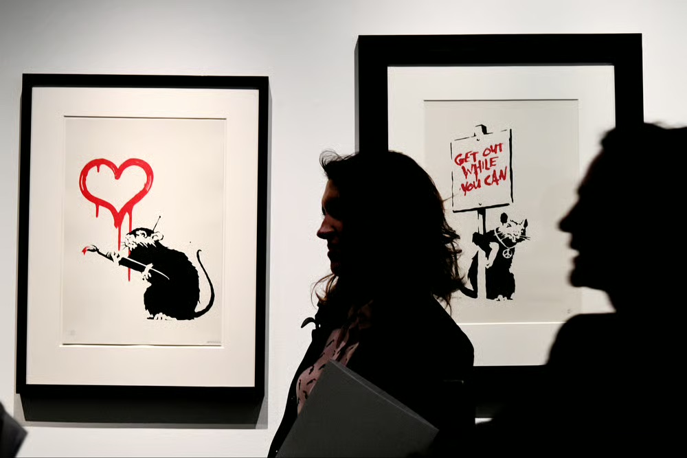 Two framed Banksy rat prints on a white gallery wall — a rat painting a dripping red heart, and a rat holding a hand-lettered red placard — with two visitors silhouetted in front of them