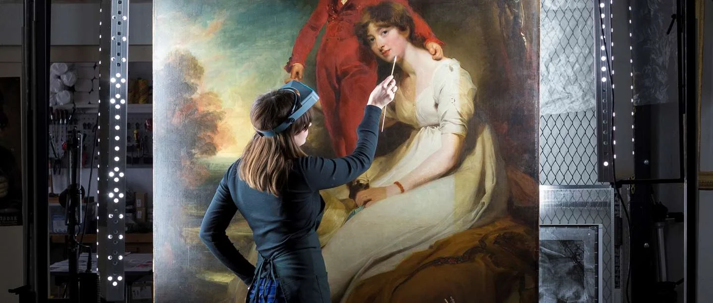 A conservator wearing a magnifying headset cleans a large portrait of a woman in a white dress, working on the canvas with a fine swab under studio lighting
