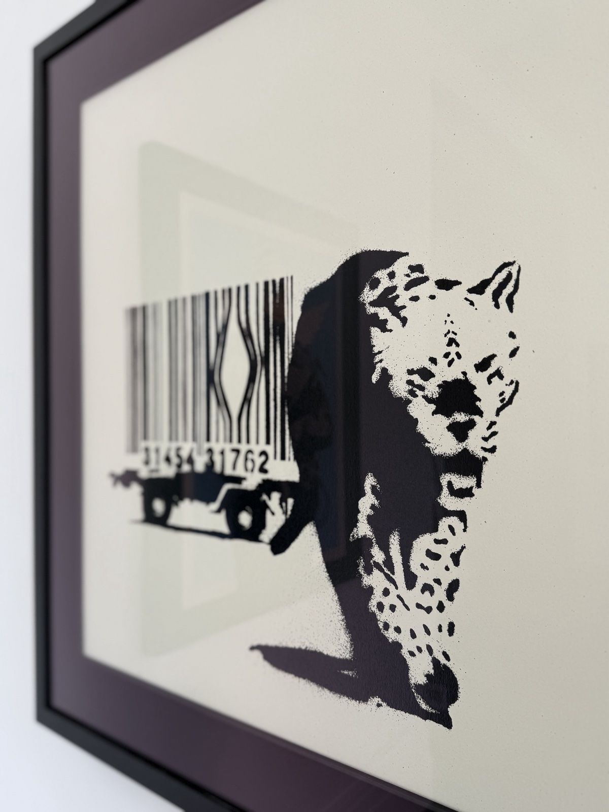 A framed print of Banksy's Barcode, a leopard stepping out of a broken barcode cage on a trailer, photographed at an angle on a white wall